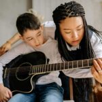 A music teacher instructs a child to play an acoustic guitar indoors.