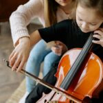 A child learning to play the cello with teacher guidance in a cozy indoor setting.