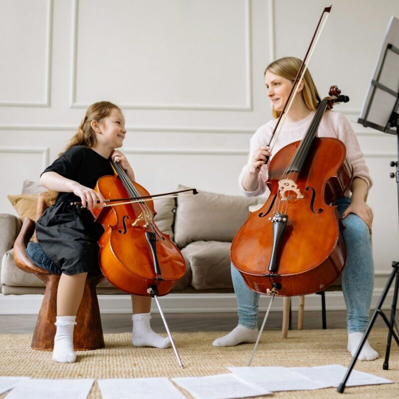 A woman and a child playing cellos together at home, emphasizing music education and bonding.